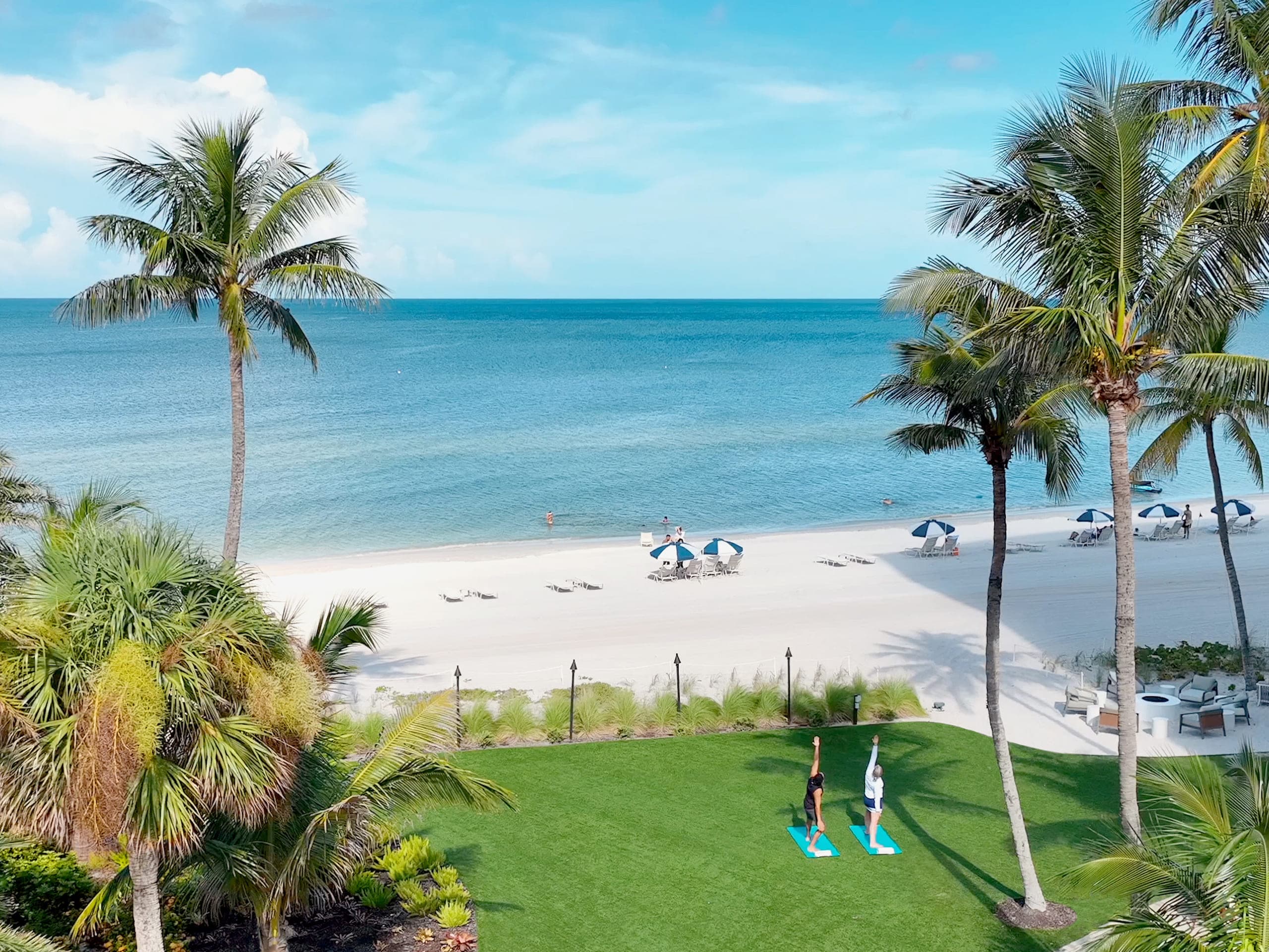 An overhead view of two people doing yoga on the lawn with the beach and water in front of them