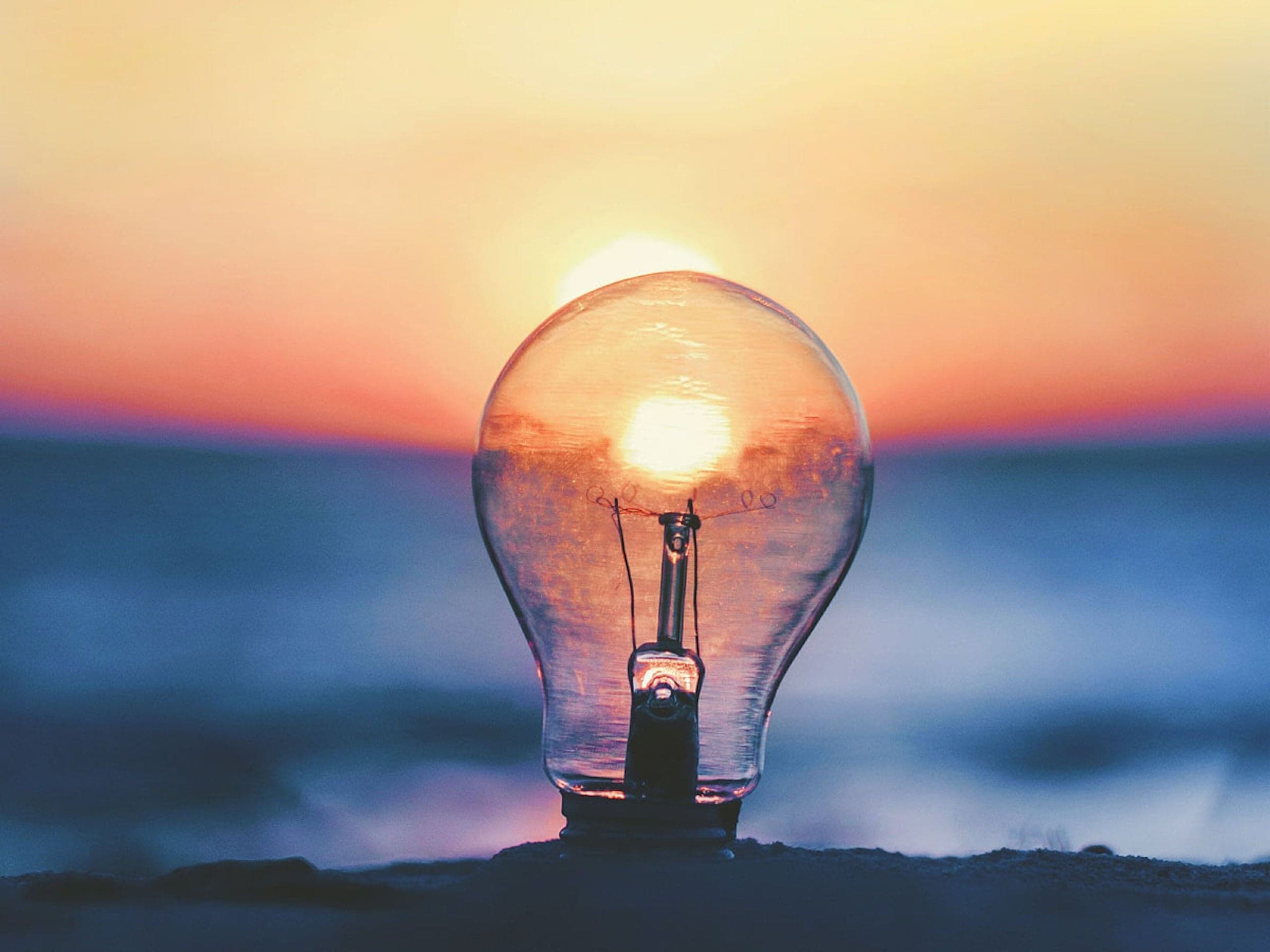 A lightbulb sitting in the sand with the sunset behind it illuminating it